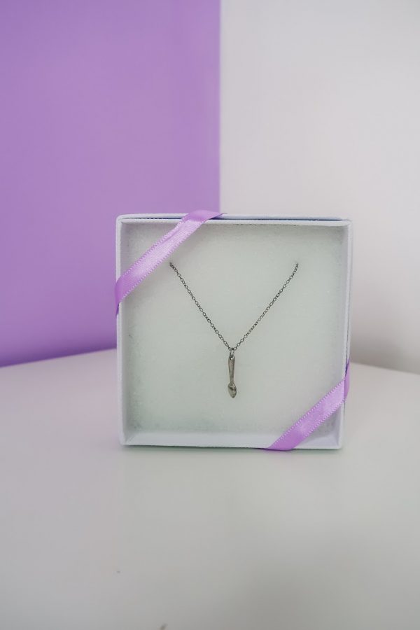 sterling silver spoonie necklace in flat white box packaging, propped up in front of lilac wall and with lilac decorative ribbon draped over opposite corners