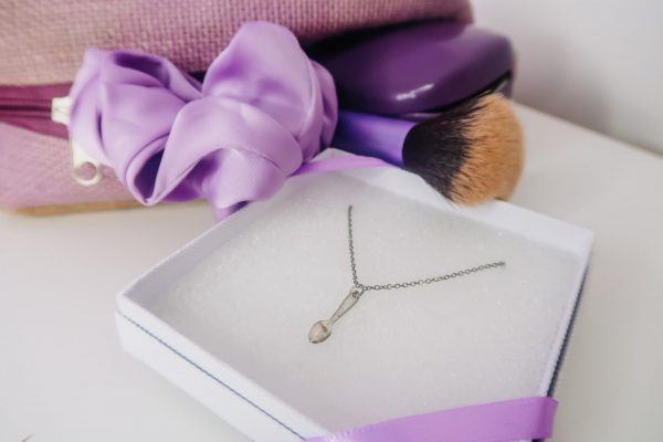flatlay of sterling silver spoonie necklace, packaged in a flat white box. lilac accessories arranged in background, including an open hessian bag, silk scrunchie, and make up brush