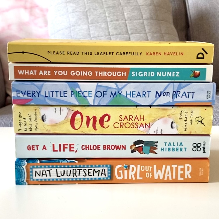 a stack of paperback books on a white table. top to bottom: please read this leaflet carefully by karen havelin, what are you going through by sigrid nunez, every little piece of my heart by non pratt, one by sarah crossan, get a life chloe brown by talia hibbert, and girl out of water by nat luurtsema