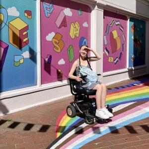 pippa in her powerchair, outdoors in the brighton lanes surrounded by colourful wall art and a rainbow on the ground. pippa has one hand up to her head shielding herself from the sun and exploring brighton by wheelchair