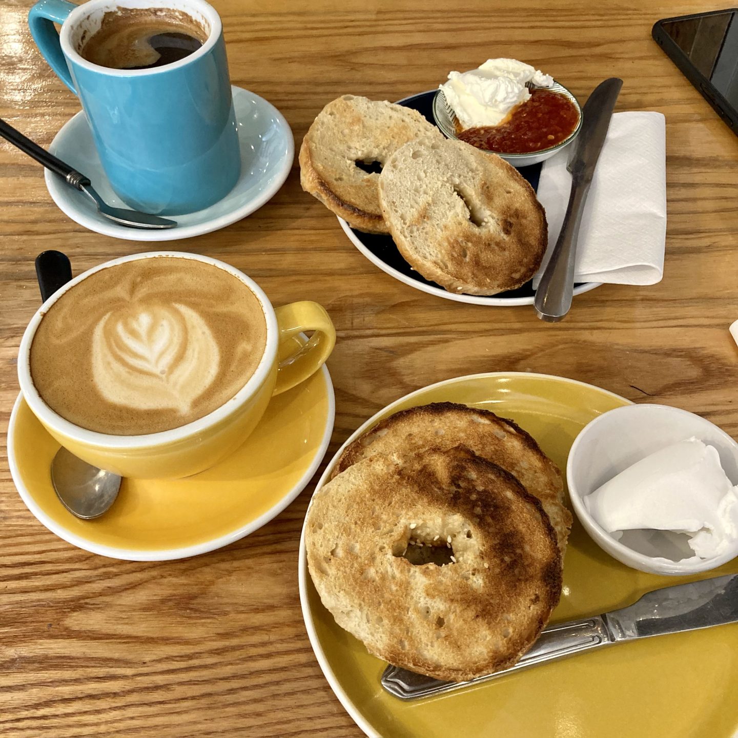 Two freshly baked bagels accompanied by vegan cream cheese and jam, alongside a latte and a cappuccino in colourful mugs