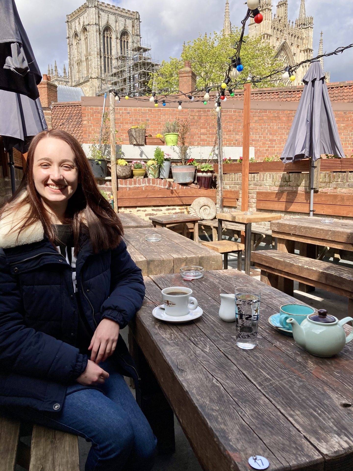 pippa sat on roof terrace at The Habit, with a cup of tea and blue teapot on the bench table in front of her and beautiful views of york minster in the background