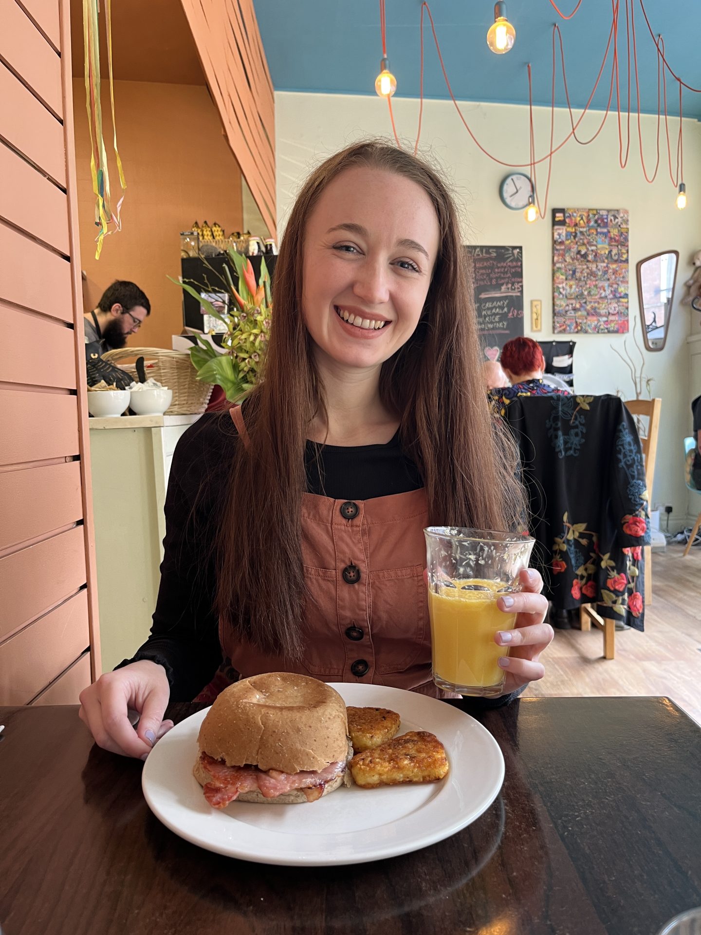 pippa sat at a table in penny's bespoke foodhouse, with a glass of fresh orange juice in one hand. in front of her is a bacon sandwich with two hash browns on the side