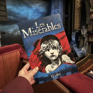 pippa's hand holding up a copy of the les mis programme in the theatre, in front of the show's backdrop