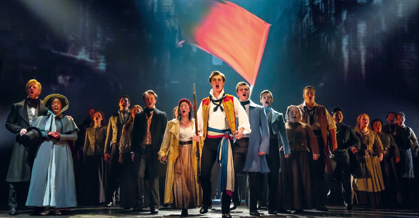 Production image of Les Mis cast stood in a row and singing, iconic red flag waving in the background.