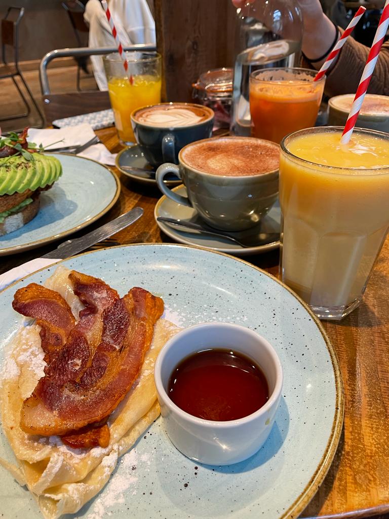 three drift in breakfasts on a table, including pancakes and avocado on toast, alongside three hot drinks and three fresh juices