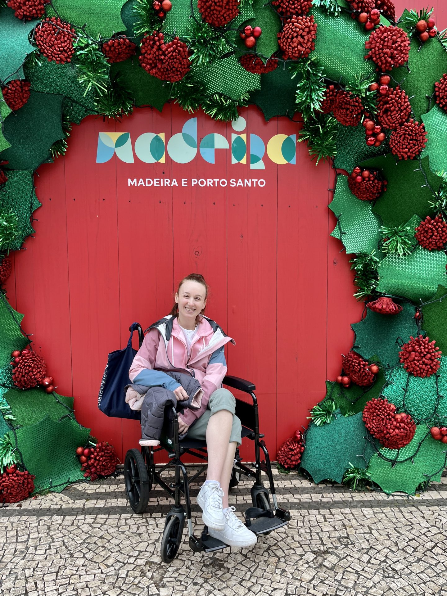 Pippa in her transit wheelchair on land in Madeira, in front of a festive photo board surrounded by a green wreath. Pippa has one leg crossed and is smiling, wearing a pink and blue raincoat and cropped khaki trousers