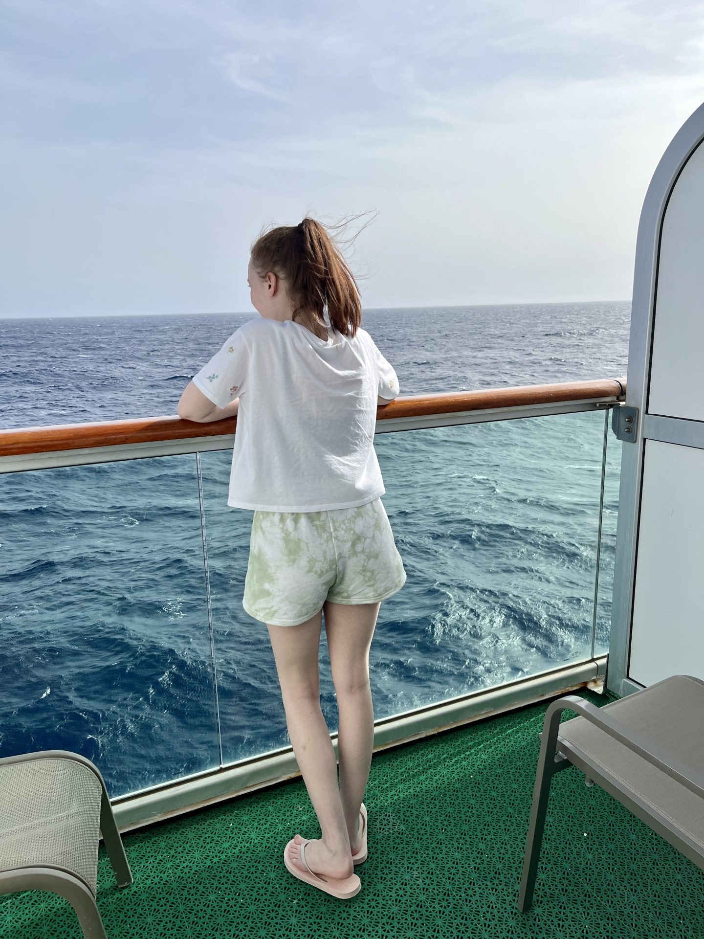 Pippa on her cabin's balcony, arms resting on the handrail and looking out to sea. She's wearing a white t-shirt, green tie dye shorts and flip flops.