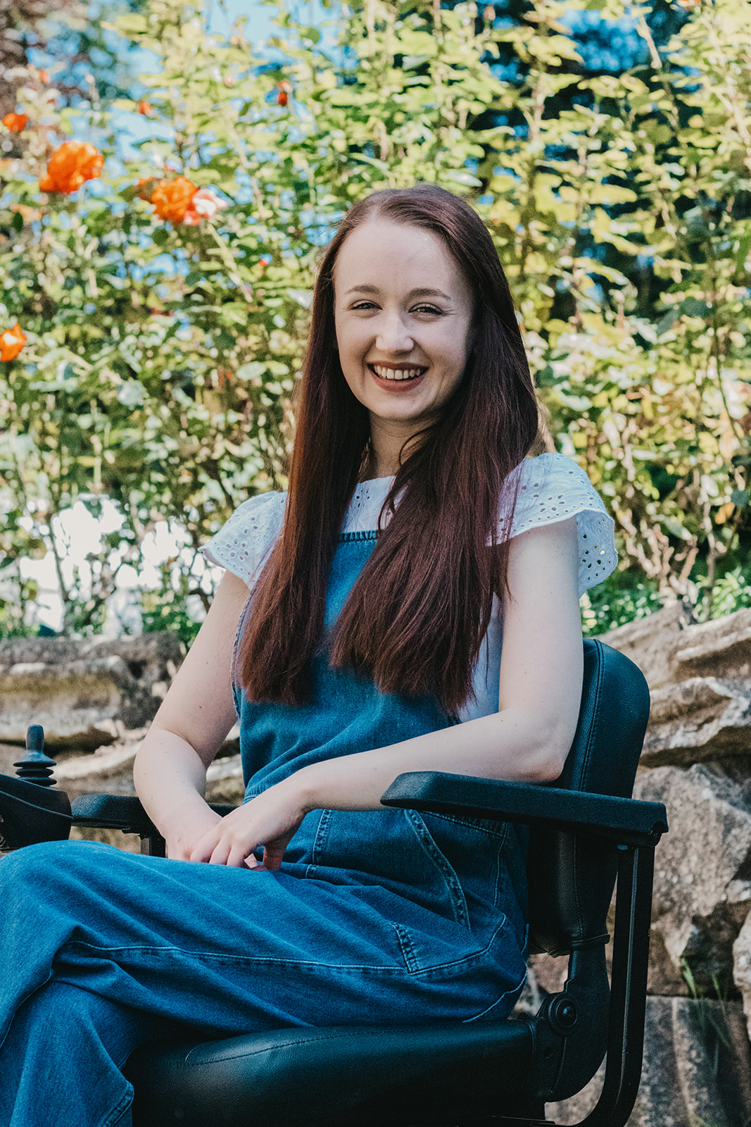 Headshot of Pippa outdoors in her power-chair, hands on her lap and smiling. She's wearing soft blue denim dungarees with a white top with floral sleeves, long brown hair down.