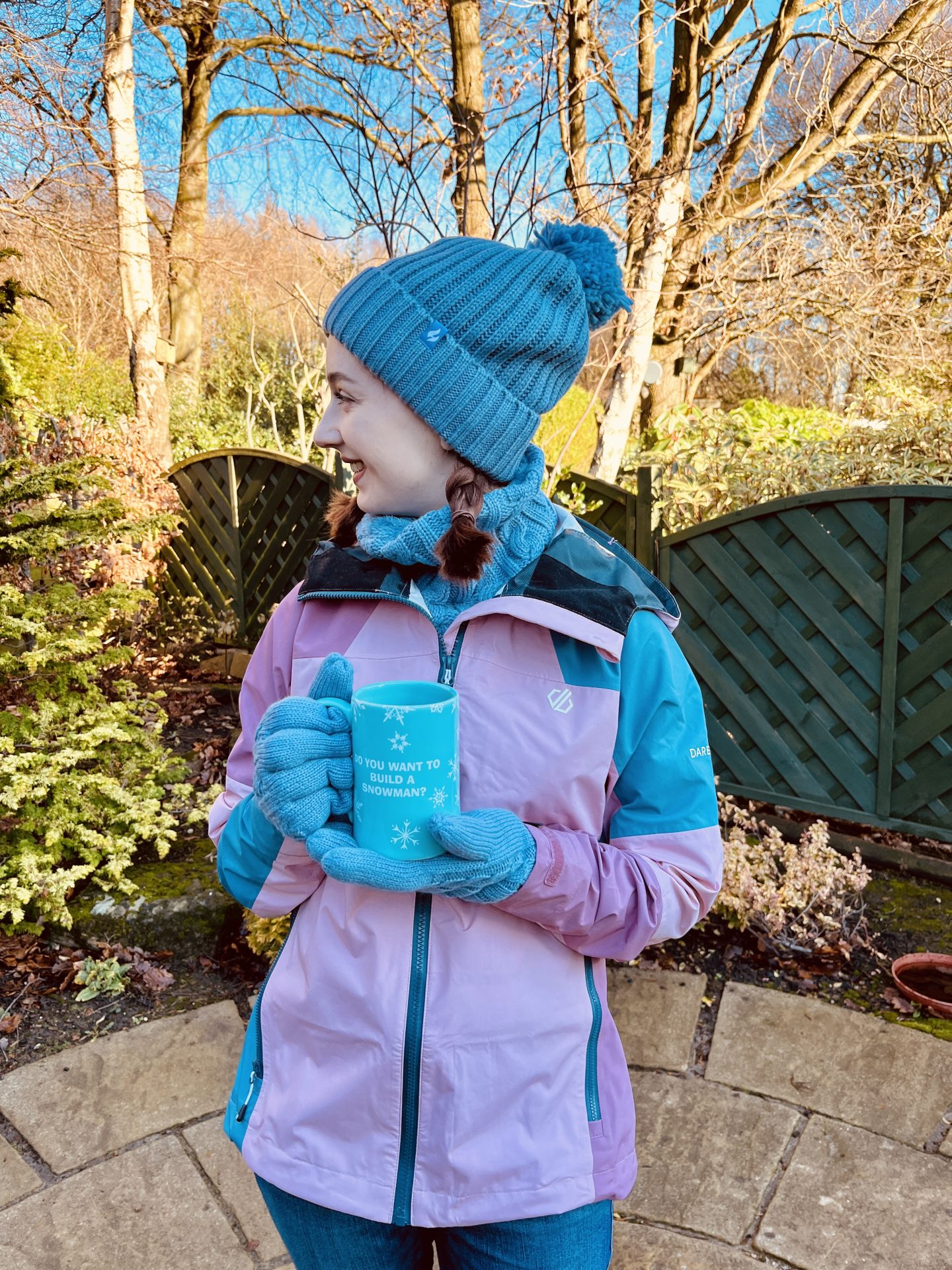 Pippa stood up outdoors, wearing her cosy new HeatHolders hat, neckwarmer and gloves in the colour dusty blue. She's also wearing a pink and blue raincoat and has her hair in plaits, looking off to the side and smiling.
