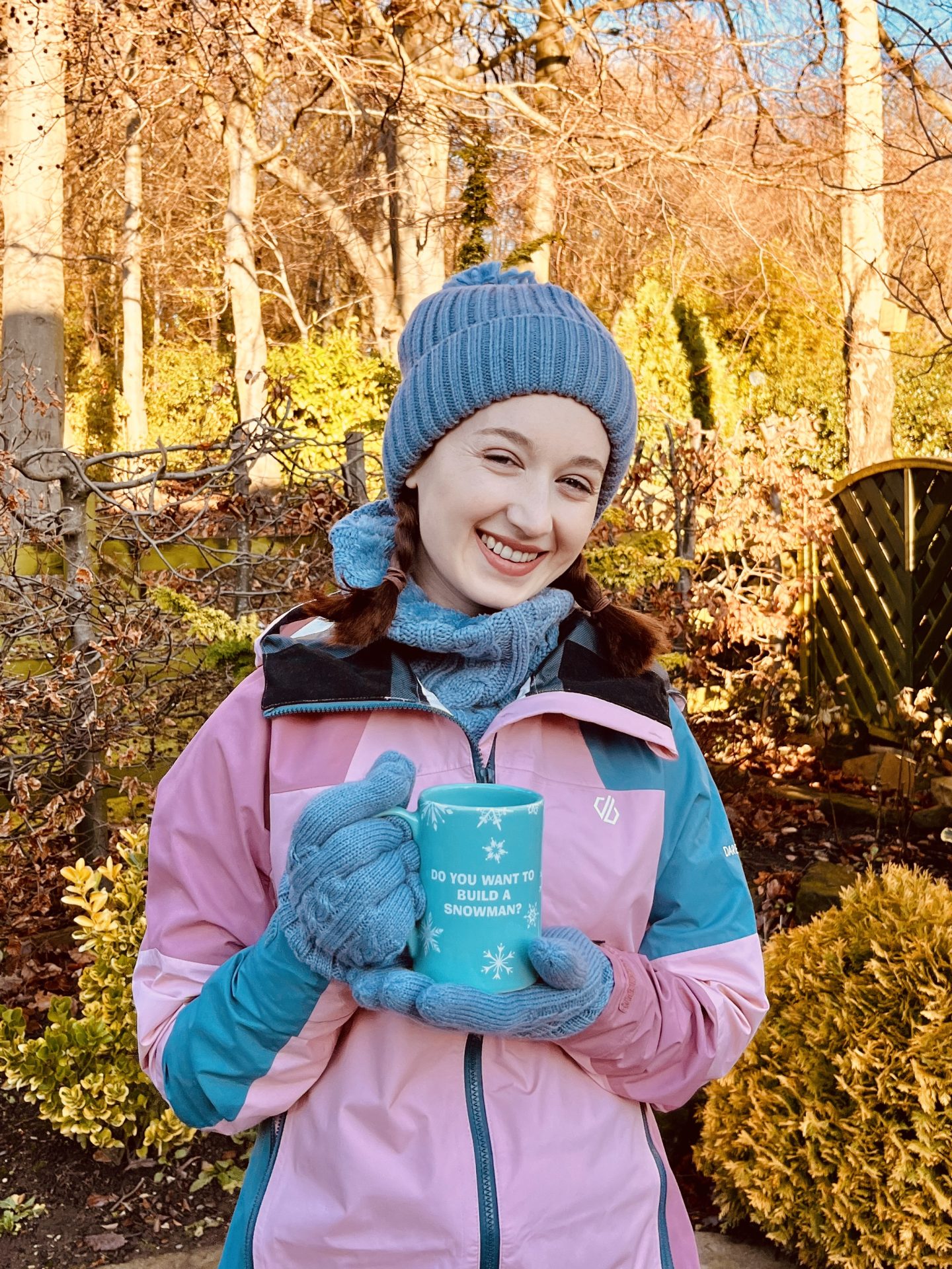 Pippa stood up outdoors, wearing her new HeatHolders hat, neckwarmer and gloves in the colour dusty blue. She's also wearing a pink and blue raincoat and has her hair in plaits, holding up a blue mug and smiling.