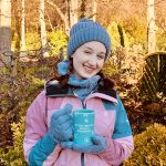 Pippa stood up outdoors, wearing her new HeatHolders hat, neckwarmer and gloves in the colour dusky blue. She's also wearing a pink and blue raincoat and has her hair in plaits, holding up a blue mug and smiling.