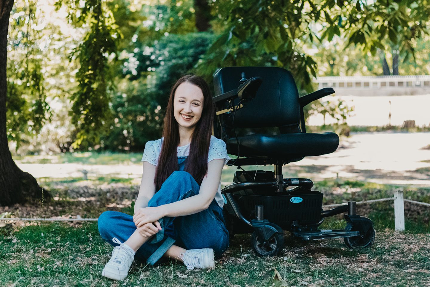 Pippa sat on the ground outdoors with her legs crossed, in front of her small black powerchair, to represent mobility aids. Pippa is smiling with her arms wrapped around her legs, wearing soft denim dungarees with a white frilly t-shirt and her long brown hair down.
