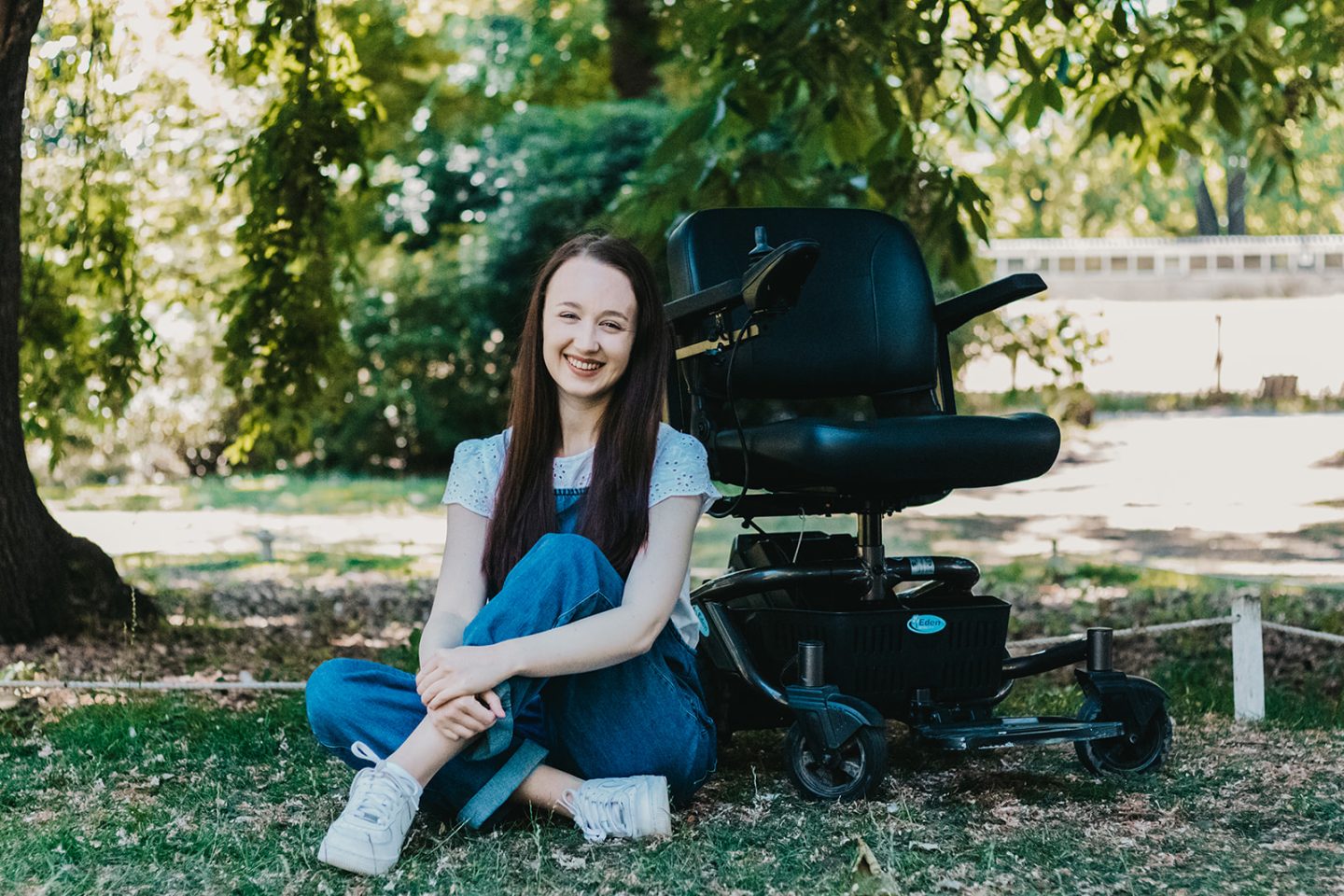 pippa sat on the ground in front of her small black powerchair, hugging one knee up to her chest and smiling. she has her long brown hair down and is wearing sift denim dungarees with a white t-shirt.