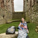 pippa in her wheelchair outside among the ruins of lindisfarne priory in northumberland. she's wearing a coat and bundle bean, and a dog backpack containing ruby, the cavapoo puppy.