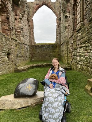 pippa in her wheelchair outside among the ruins of lindisfarne priory in northumberland. she's wearing a coat and bundle bean, and a dog backpack containing ruby, the cavapoo puppy.