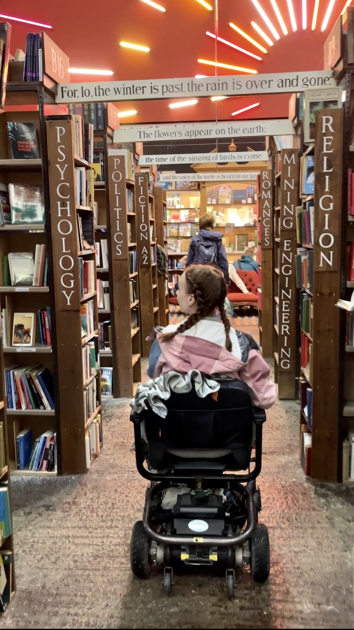 Pippa in her wheelchair inside Barter Books in Northumberland, making her way down aisles of bookshelves and pausing to look at the psychology section.