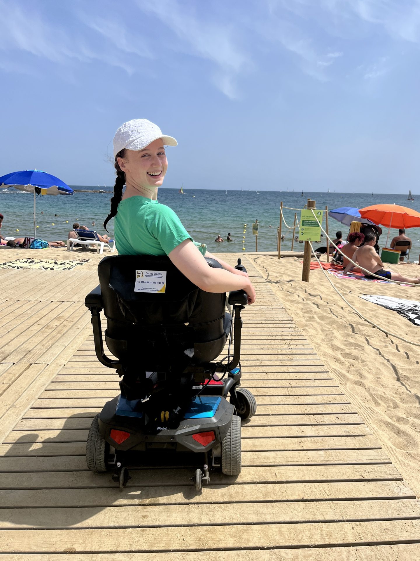 Pippa in her wheelchair on a wooden walkway at Nova Icaria Beach, turning back to look at the camera with the sea visible behind her. A highlight of wheelchair accessible Barcelona.