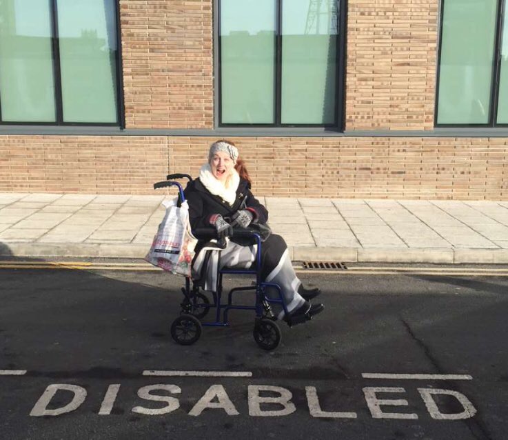 Pippa in her transit wheelchair, one of her everyday mobility aids, laughing and pointing down at the word 'disabled' printed on the road on the accessible parking space that she's in.