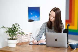 Pippa sat at a desk with her laptop open in front of her, looking down and writing in a diary to represent freelancing with a chronic illness. Professional image by Timm Cleasby Photography.