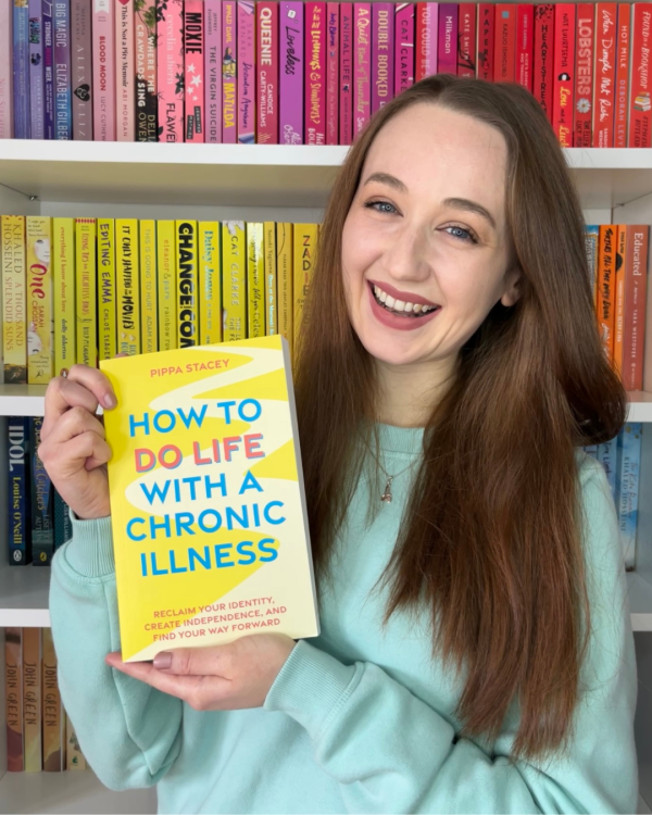 pippa holding up her book, how to do life with a chronic illness, in front of her rainbow bookcase and smiling