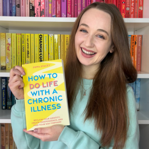 Pippa stood in front of her rainbow bookcase, smiling and holding up a copy of her book, How To Do Life With A Chronic Illness. The paperback book is yellow with blue and orange typography and a winding path design in the background.