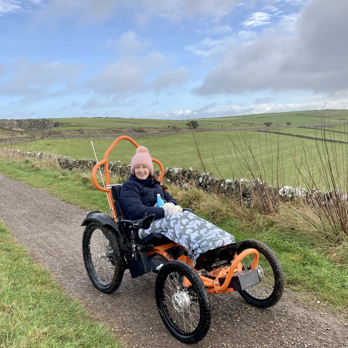 Pippa in the Boma 7 off-road wheelchair, with her legs elevated in front of her and with beautoful green Derbyshire countryside in the background. Pippa is wearing a hat, coat and gloves, holding a water bottle and wearing a wheelchair cosy over her legs.