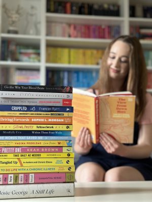 A colourful stack of non-fiction books by disabled authors, with Pippa sat on her sofa reading a book in the background.