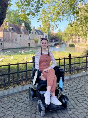 Pippa in her wheelchair in Minnewater park in Bruges, with the river and houses lining the water behind her. Pippa is smiling with her hands in her lap, wearing burnt orange dungarees with a stripy top.