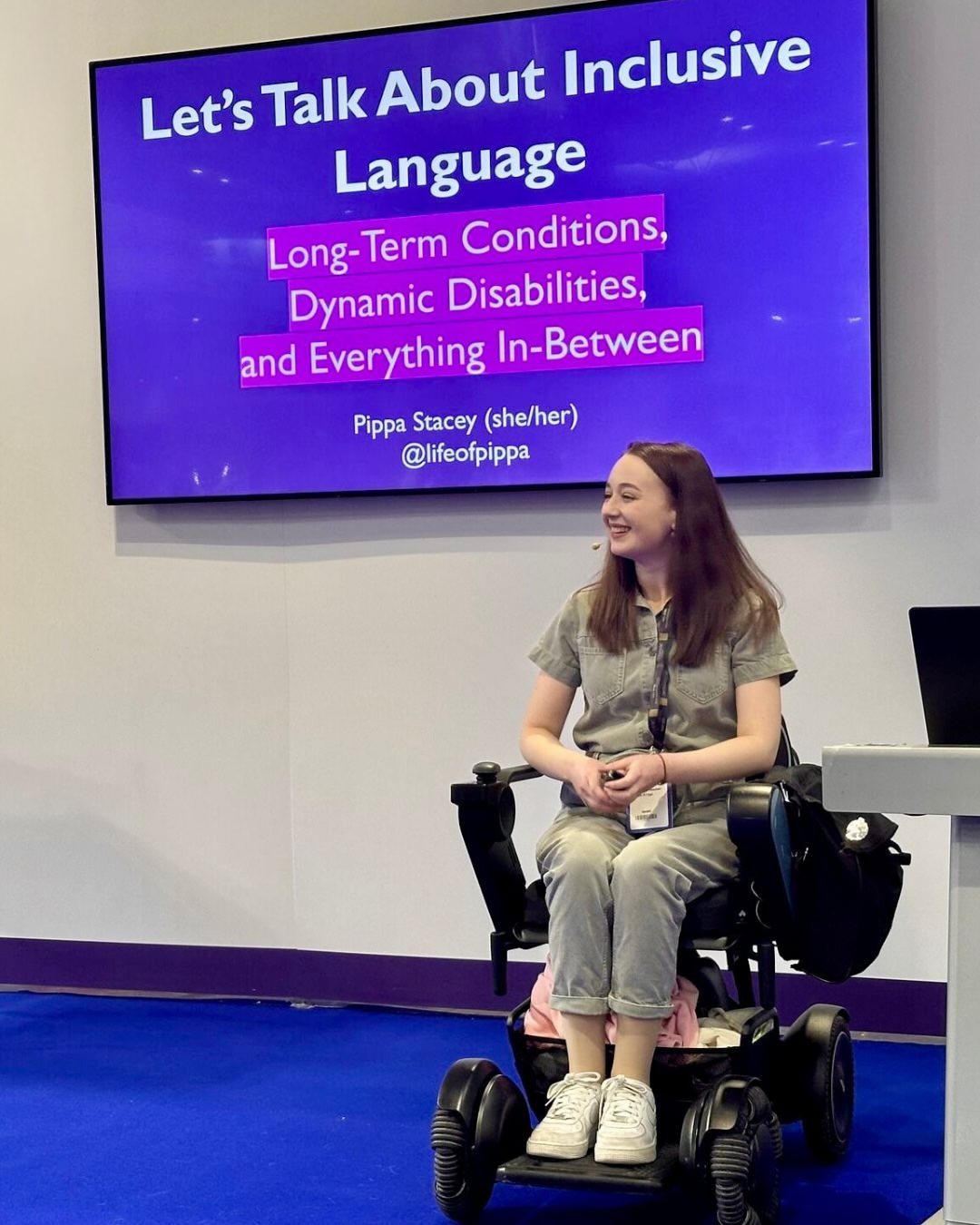 Pippa in her wheelchair presenting at Naidex, wearing a khaki jumpsuit and smiling at the crowd. The screen behind her shows the title page of her 'let's talk about inclusive language' session.