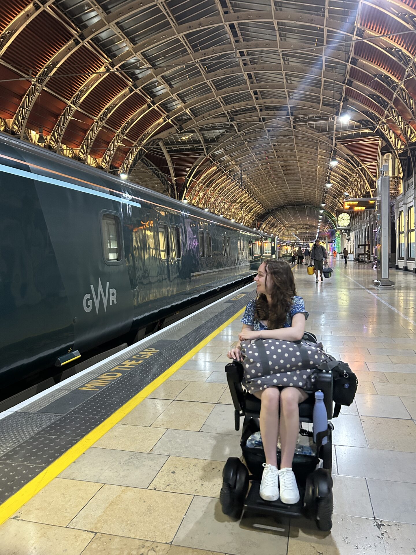 Pippa on the platform at London Paddington, looking at the sleeper train and getting ready to board as a wheelchair user. She has a dotty overnight bag on her knee.