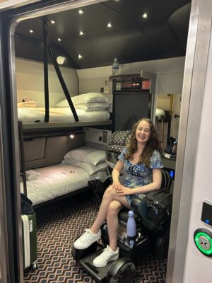 Pippa, a white female wheelchair user, wearing a blue dress and in the accessible cabin on board the Night Riviera Sleeper Train.
