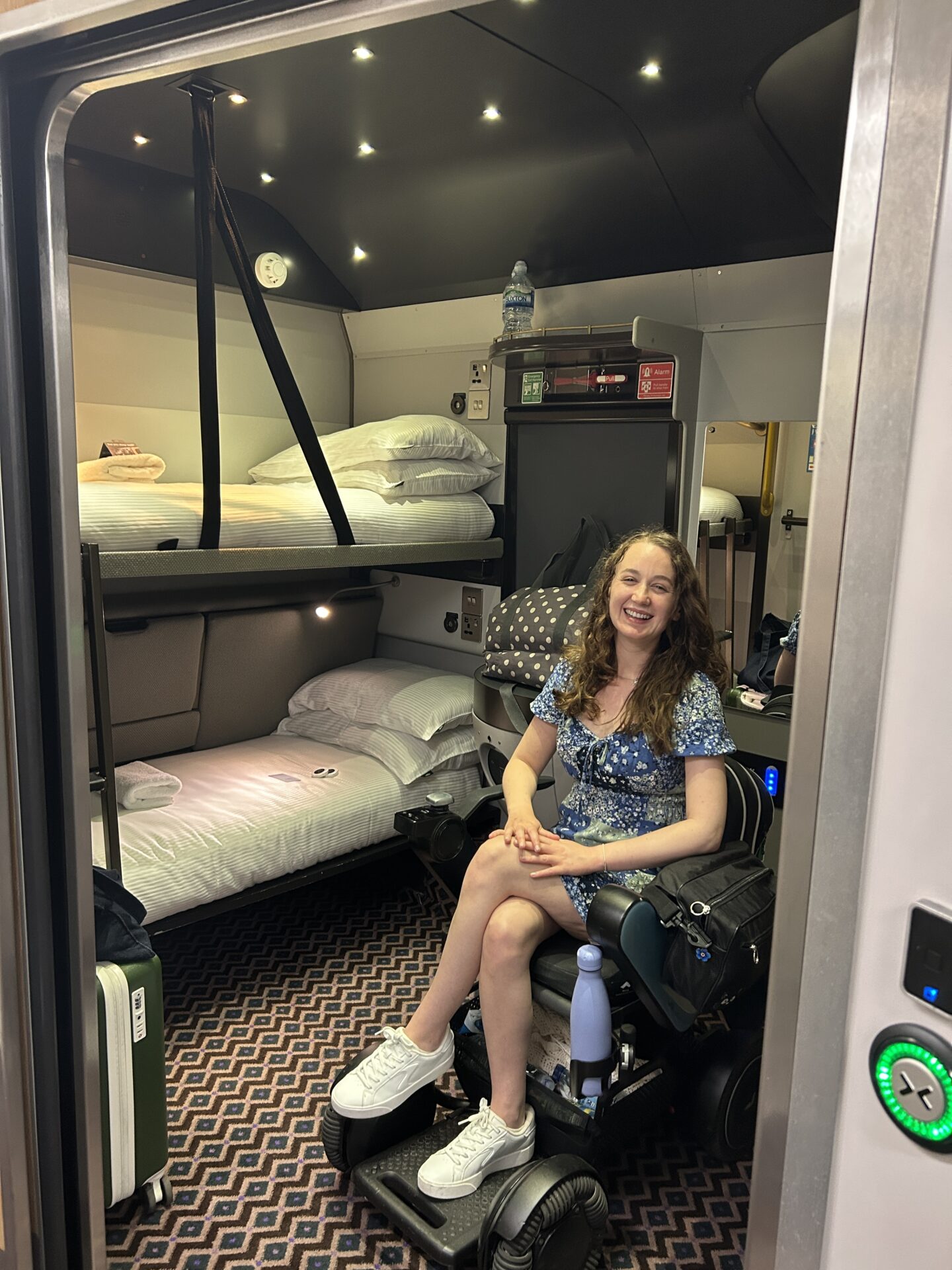 Pippa in the accessible cabin on the Sleeper Train, in her wheelchair. In the cabin are two bunk beds with clean towels, a sink, a mirror, and a small window with a blind pulled down.