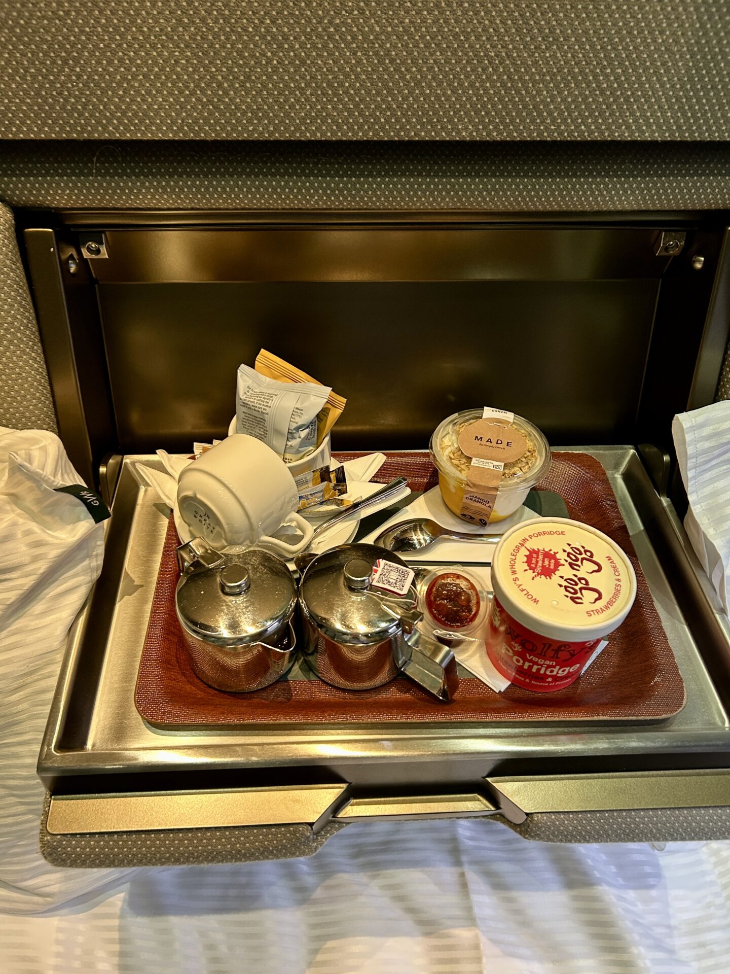 Breakfast on a silver tray on the sleeper train. Includes pots of tea and coffee, a pot of granola, a tub of ready made porridge, and biscuits and cutlery.