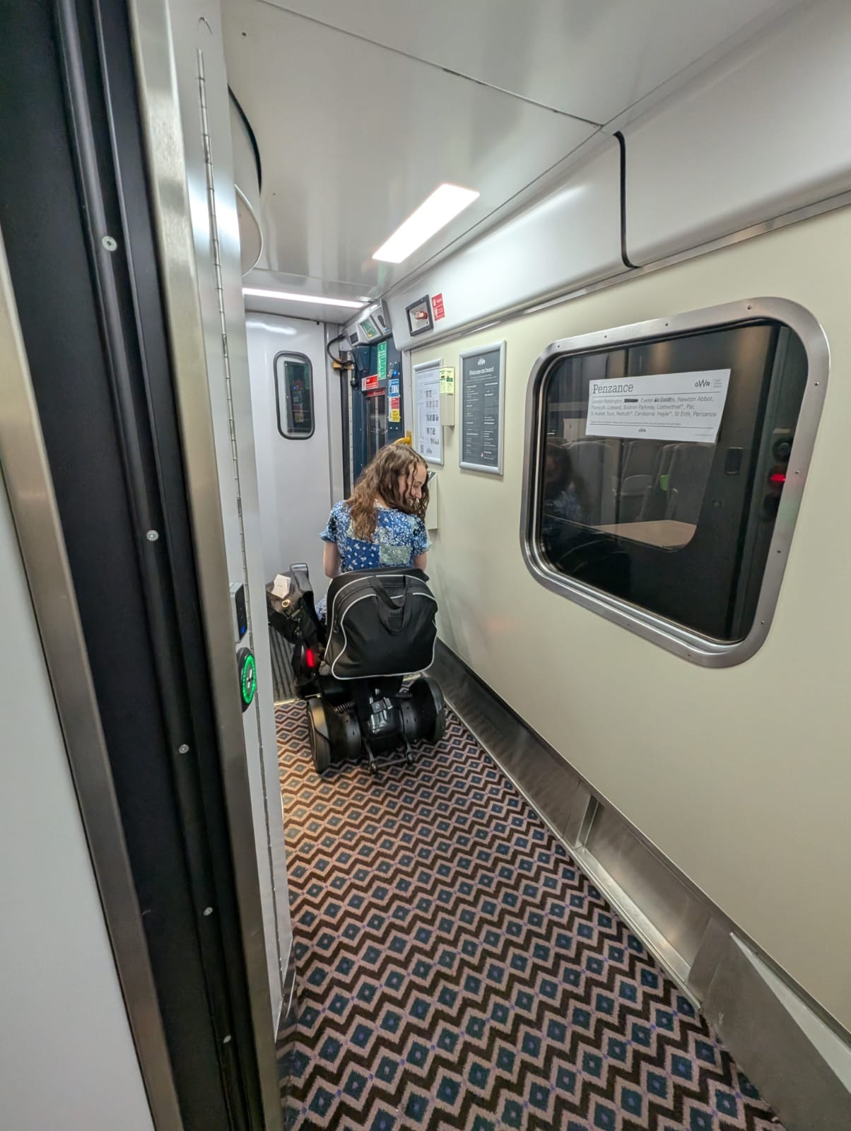 Pippa in the sleeper train corridor using her wheelchair, back to the camera and looking down. Sign on the window reads 'Penzance', followed by a list of stops.